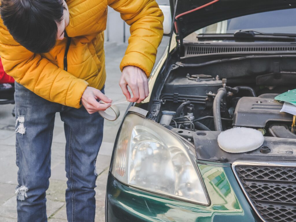 A young caucasian guy in a yellow jacket sticks scoich around the edges of the headlight preparing i